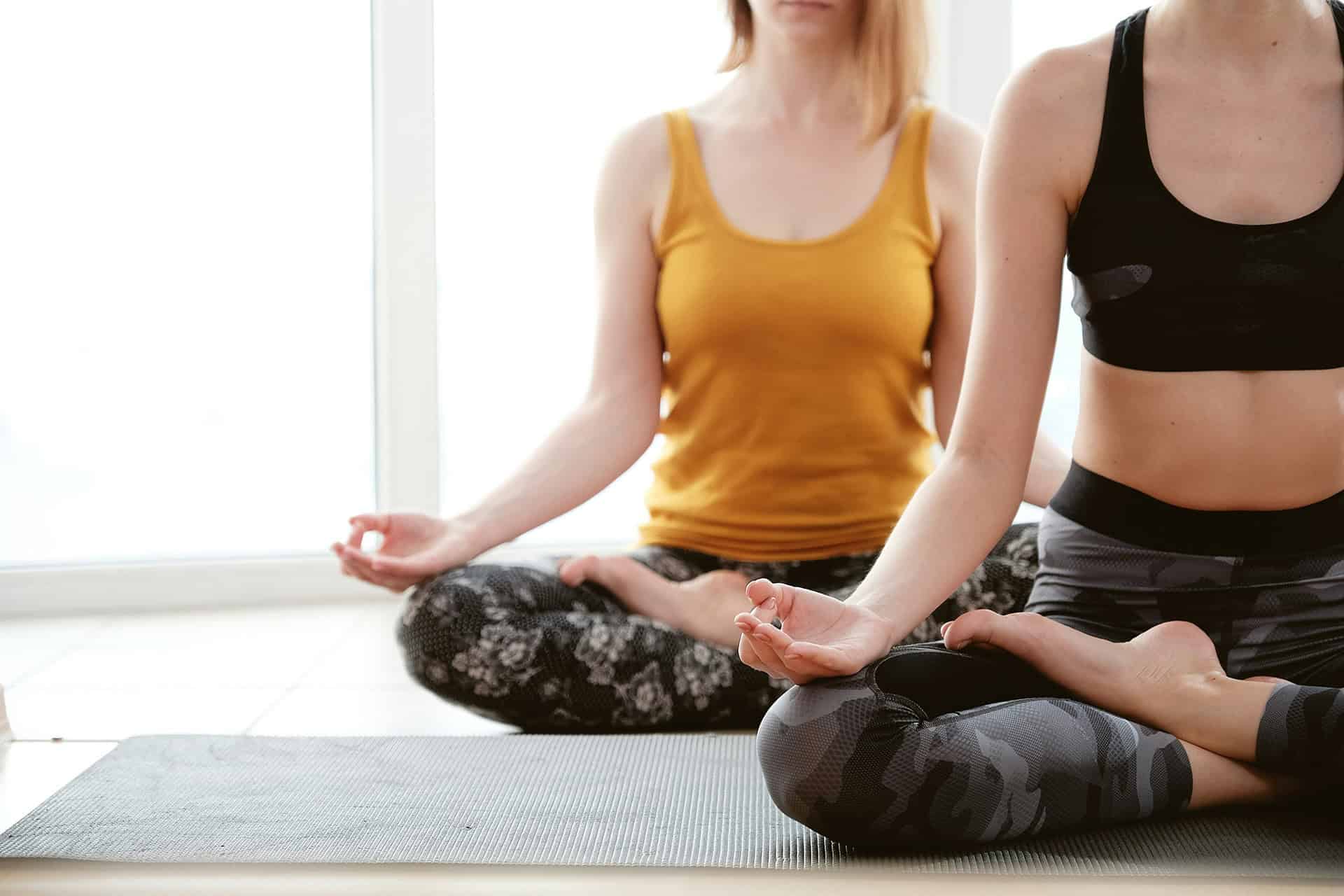 Two women meditate in lotus position on a yoga mat in front of a window.